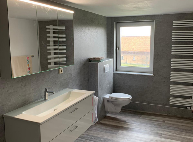 Modernes Badezimmer mit fugenlosen Wänden, wandhängender Toilette und großzügigem Waschtisch. Dezente Grau- und Holztöne verleihen dem Raum eine elegante und zeitlose Optik.