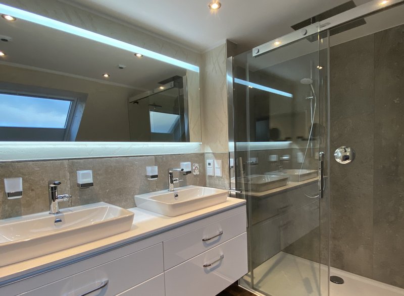 Modernes Badezimmer mit Doppelwaschtisch, beleuchtetem Spiegel und bodengleicher Regendusche. Hochwertige Materialien und eine elegante Glasabtrennung sorgen für ein luxuriöses Ambiente.